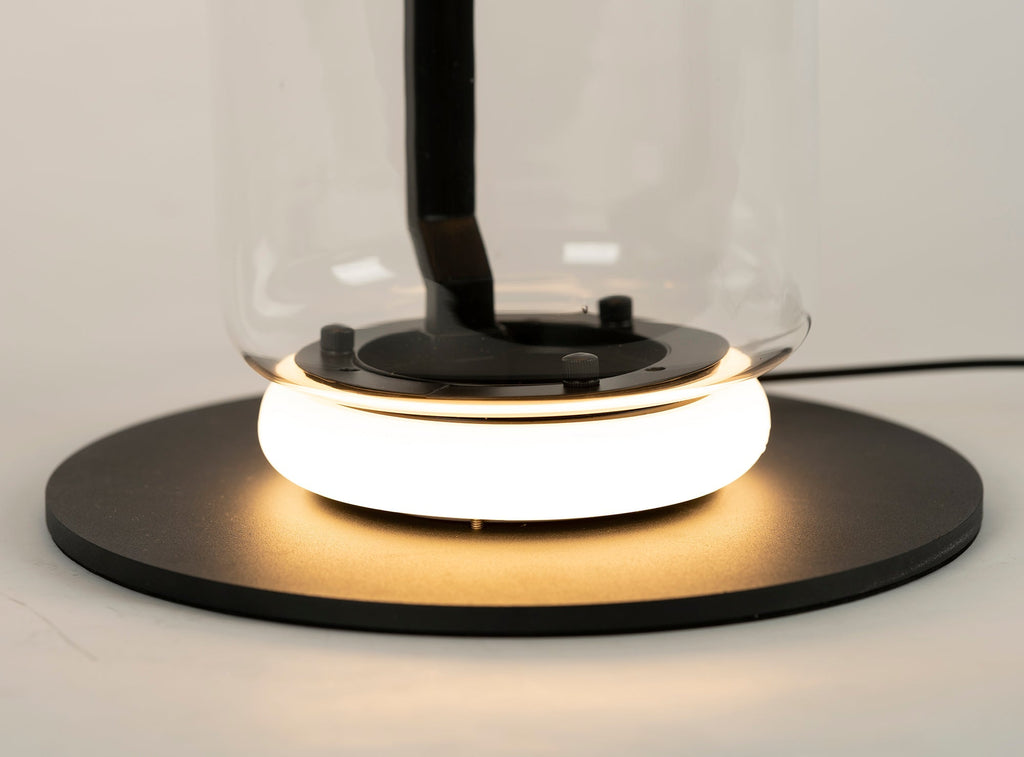 Close view of the glowing ring and black base plate at the foot of this Clérachic floor lamp, with the glass cylinder rising above.