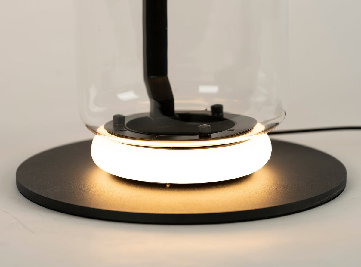 Close view of the glowing ring and black base plate at the foot of this Clérachic floor lamp, with the glass cylinder rising above.