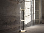 Clérachic floor lamp with a flared glass top and two cylinders, lit in a worn plaster room with a tall door behind.