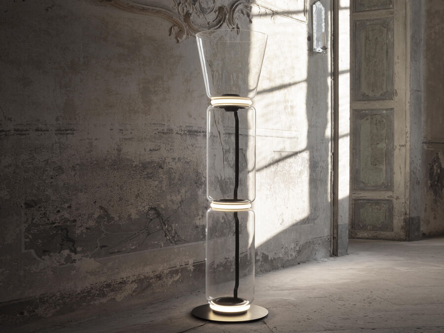 Clérachic floor lamp with a flared glass top and two cylinders, lit in a worn plaster room with a tall door behind.