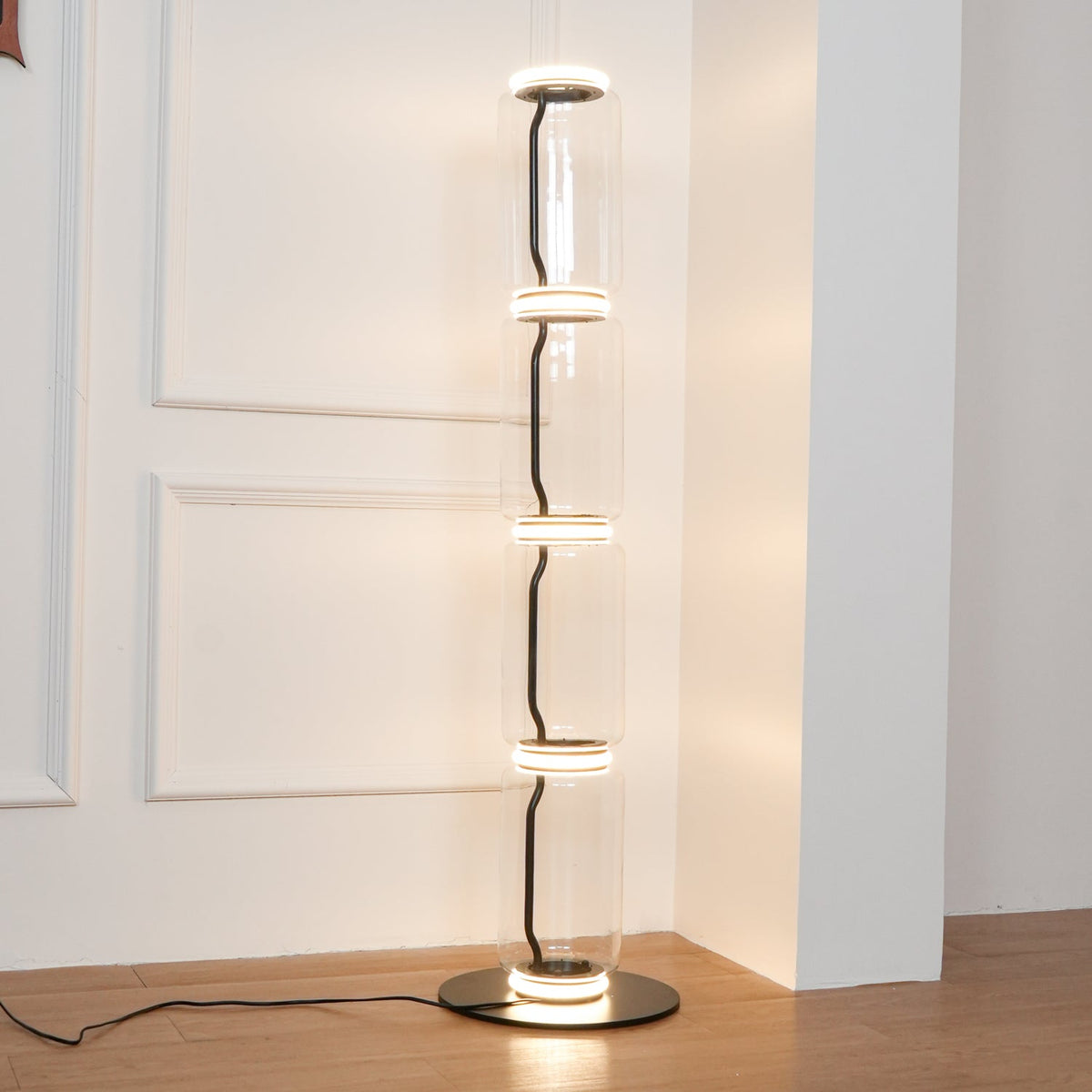 Clérachic floor lamp lit in a white panelled corner, a black cord curling between its four glass cylinders, on a wood floor.