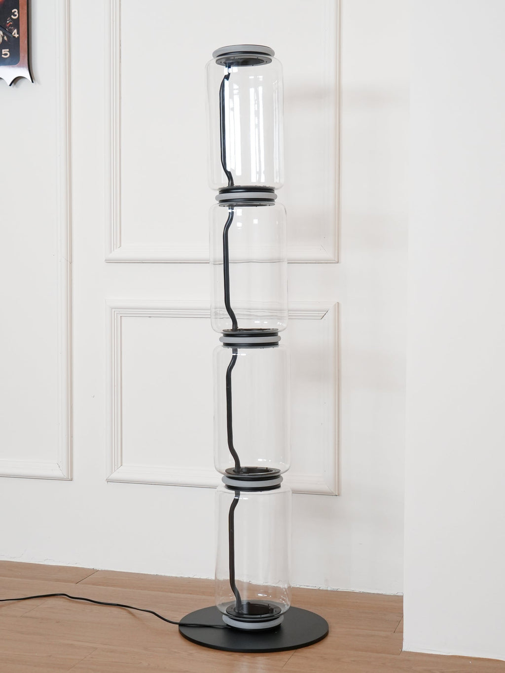 Unlit Clérachic floor lamp on a round black base in a white panelled corner, four glass cylinders joined by dark metal collars.