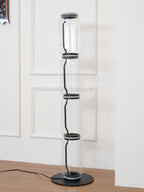 Unlit Clérachic floor lamp on a round black base in a white panelled corner, four glass cylinders joined by dark metal collars.