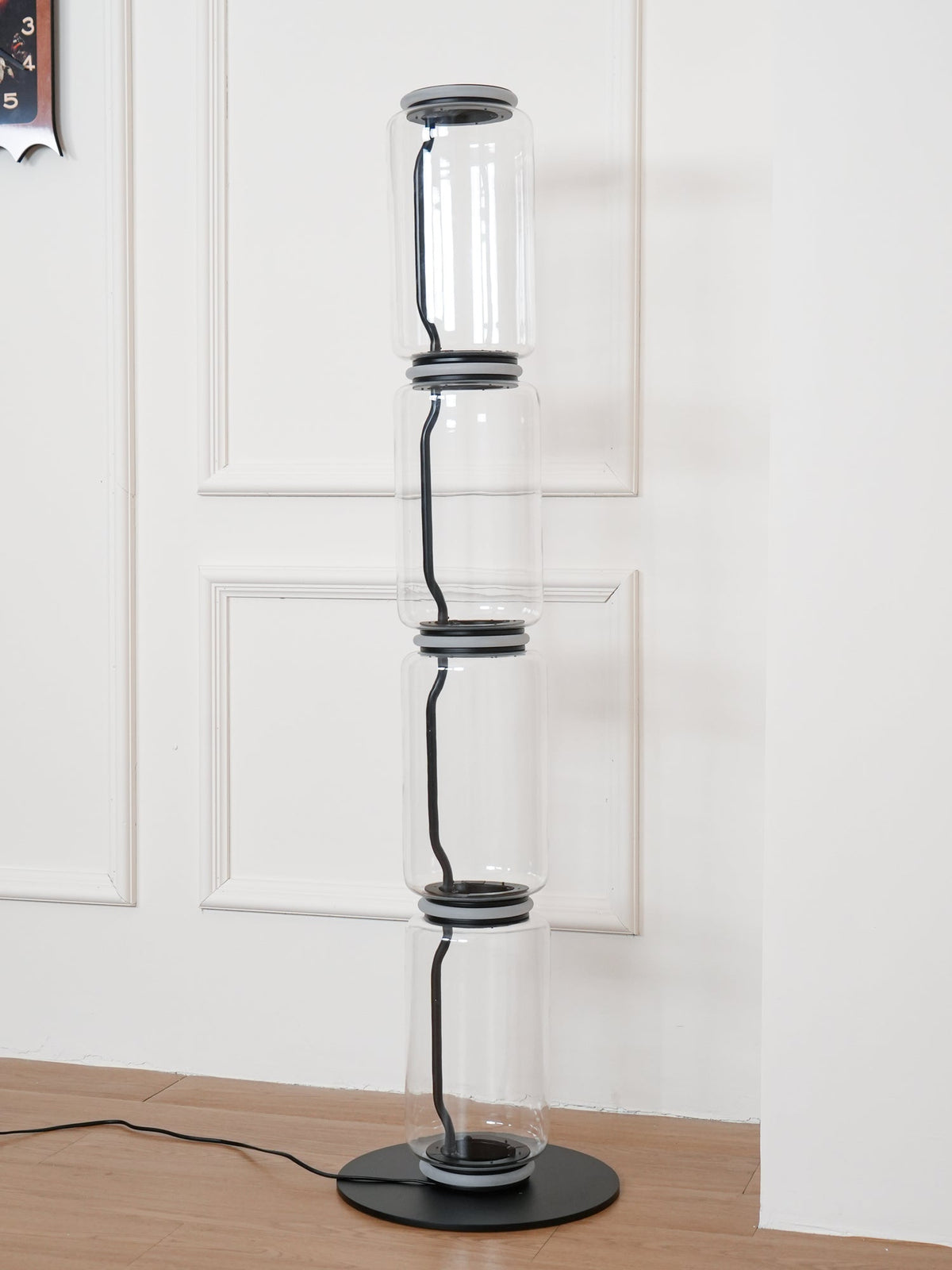 Unlit Clérachic floor lamp on a round black base in a white panelled corner, four glass cylinders joined by dark metal collars.