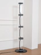 Unlit Clérachic floor lamp of four glass cylinders in a white corner, on a wood floor with a black cord at the base.
