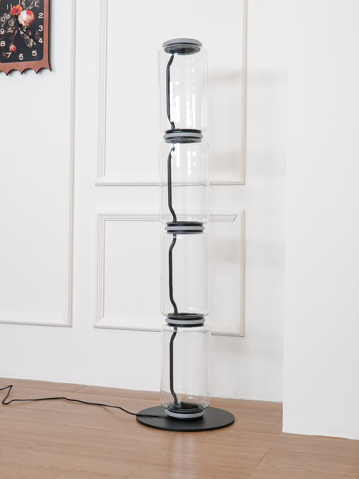 Narrow view of an unlit Clérachic floor lamp on a round black base, in front of a white panelled wall and a small framed picture.