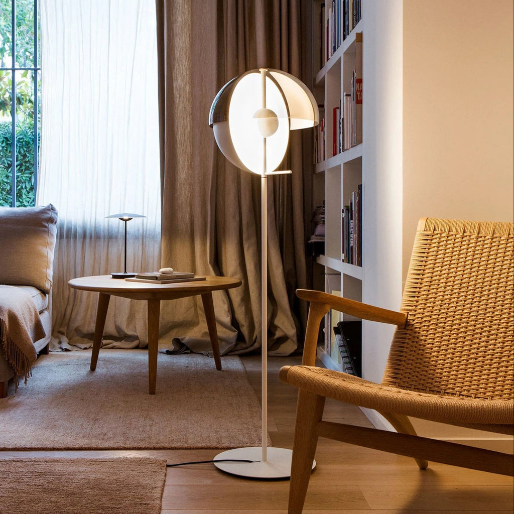 This Clérachic floor lamp stands with its white stem and open dome shade beside a woven armchair, a low round table and tall curtains.