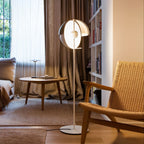 This Clérachic floor lamp stands with its white stem and open dome shade beside a woven armchair, a low round table and tall curtains.