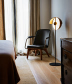 Black Clérachic floor lamp with its dome tilted open beside a leather armchair, a bed and a dark wooden chest of drawers.
