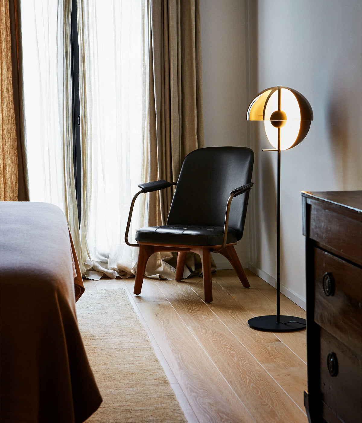 Black Clérachic floor lamp with its dome tilted open beside a leather armchair, a bed and a dark wooden chest of drawers.