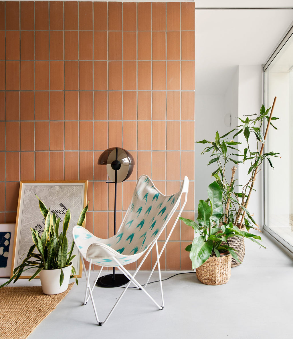 This Clérachic floor lamp stands against an orange-tiled wall, beside a butterfly chair, a framed print and leafy plants.