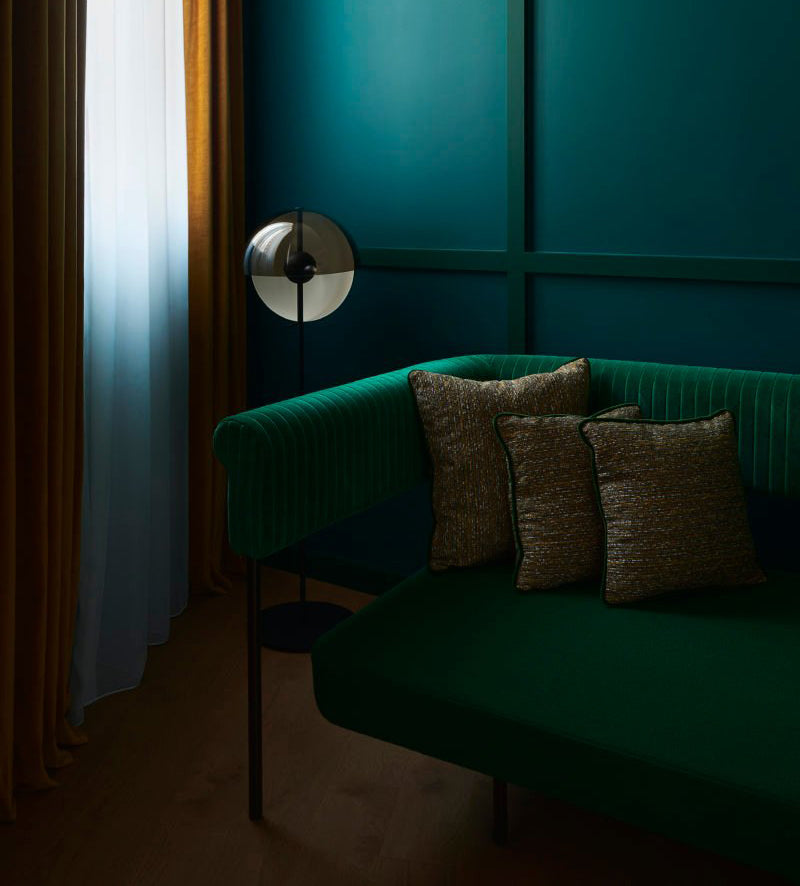 Floor lamp with its dome tilted down, in front of a deep teal wall and a green velvet sofa with patterned cushions; Clérachic.