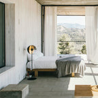 A Clérachic floor lamp stands beside a low bed with a grey blanket, in a bedroom with sheer curtains and a view of the hills.