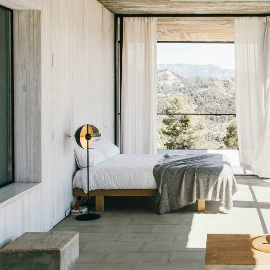 A Clérachic floor lamp stands beside a low bed with a grey blanket, in a bedroom with sheer curtains and a view of the hills.
