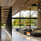 This Clérachic floor lamp glows on the right of an open-plan lounge with a grey sofa, a timber ceiling and glass walls.
