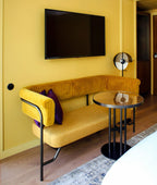 Small Clérachic floor lamp beside a mustard velvet sofa and a round glass side table, on a yellow wall below a wall-mounted television.
