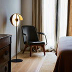 Black Clérachic floor lamp with its open dome lit, beside a leather armchair and floor-length curtains in a calm bedroom.
