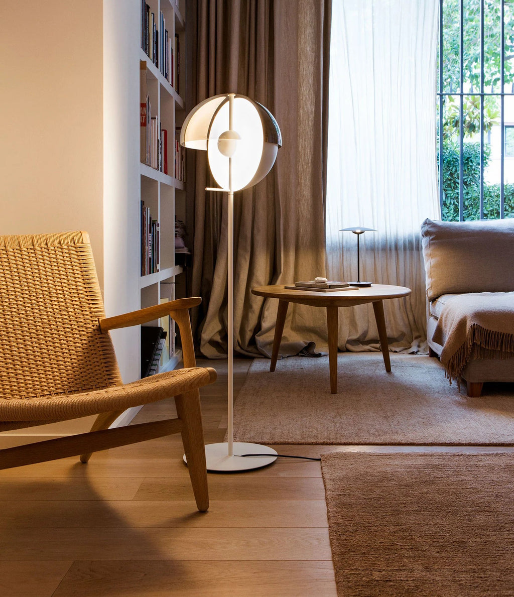White-stemmed Clérachic floor lamp with a lit open dome, beside a woven armchair and a low round table in front of shelves.