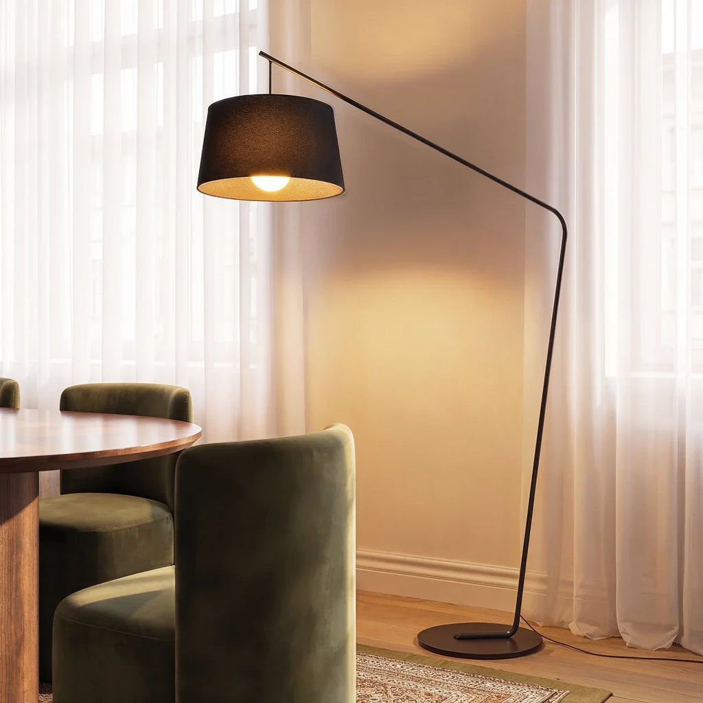 Clérachic black arc floor lamp with a tapered fabric shade glowing beside sheer curtains and a green velvet dining chair.