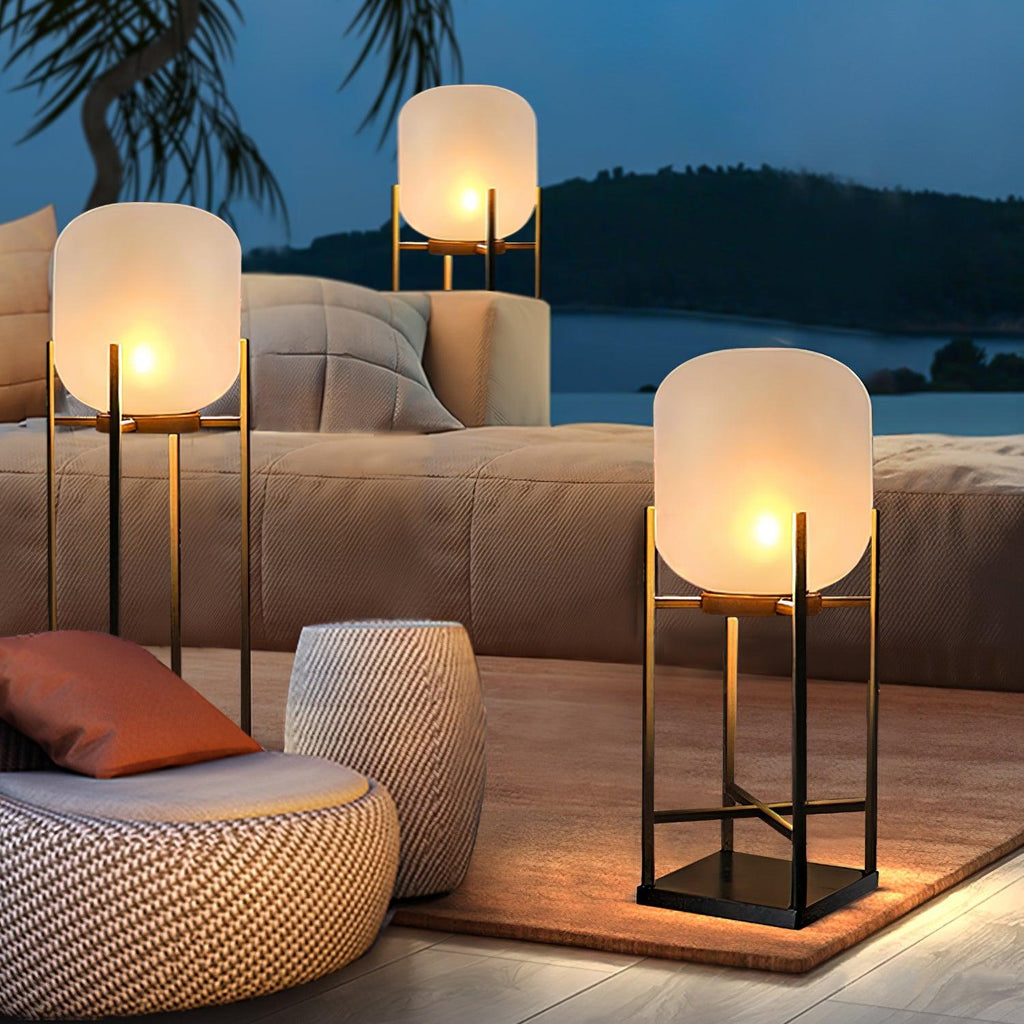 Three lit Clérachic frosted glass lanterns of different heights on a patio rug by a sofa, with a lake in the dusk behind.