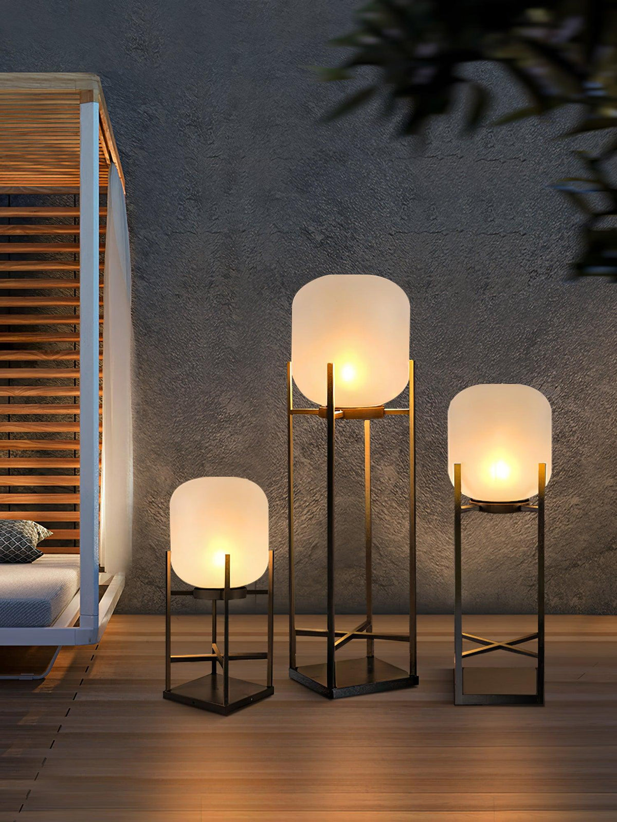 Three Clérachic frosted glass lanterns in graduated heights glowing on a deck beside a grey wall and a slatted pergola.