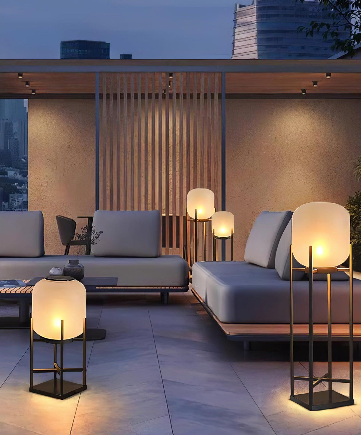 Clérachic lanterns glowing on a paved terrace by a grey outdoor sofa, with a slatted screen and city towers behind.