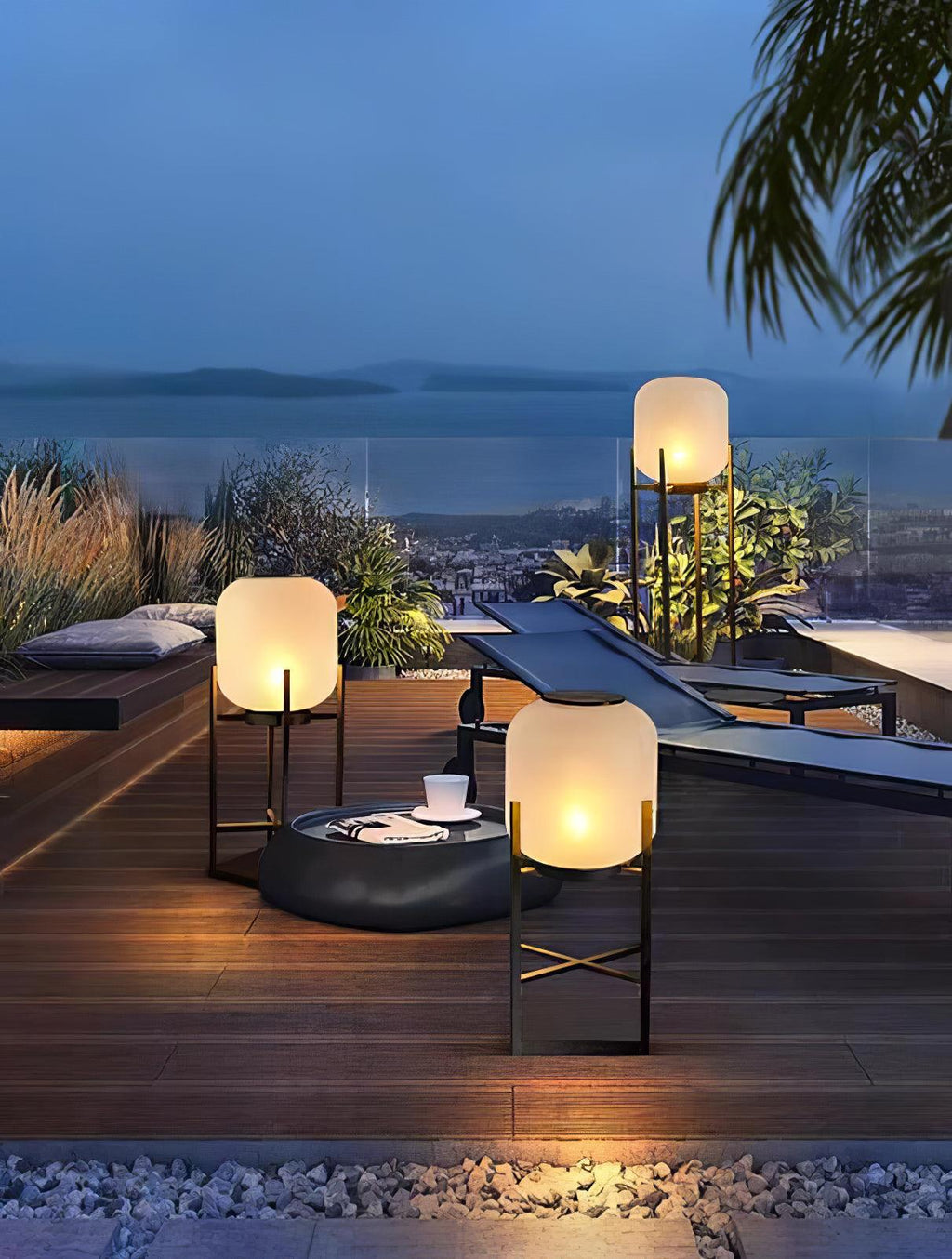 Three lit Clérachic lanterns on a roof terrace deck with a round fire bowl, with a sea view in the blue dusk.