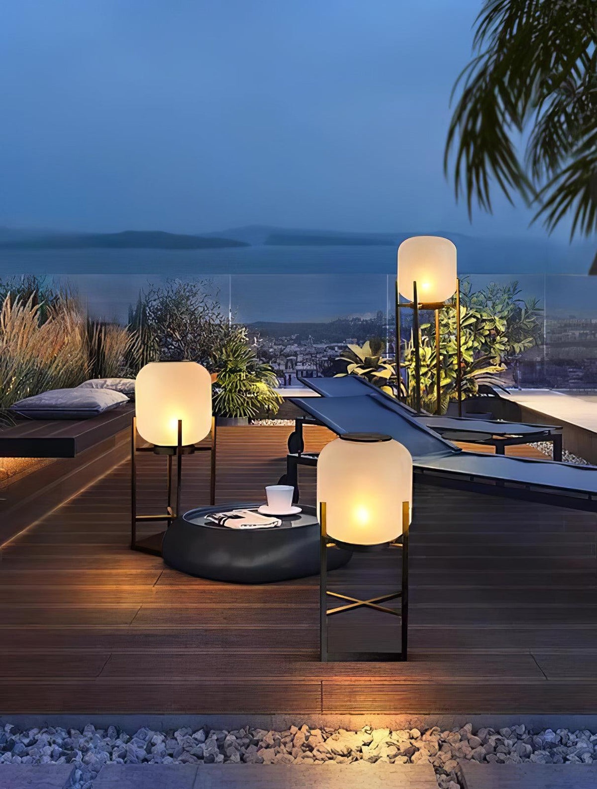 Three lit Clérachic lanterns on a roof terrace deck with a round fire bowl, with a sea view in the blue dusk.