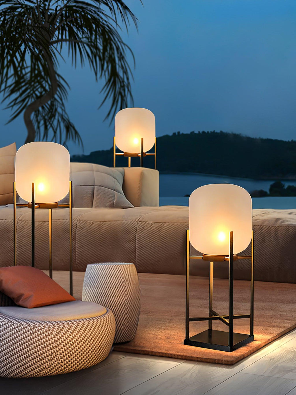 Three Clérachic lanterns glowing beside a taupe outdoor sofa and a woven pouffe, with the sea behind in blue dusk light.