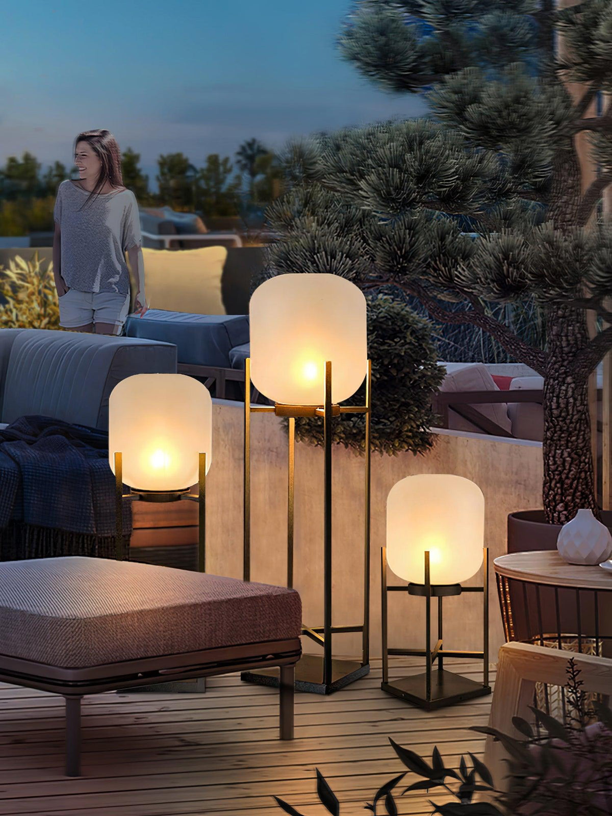 Clérachic lanterns glowing on a terrace at dusk, with a woman standing among pines and an outdoor sofa nearby.