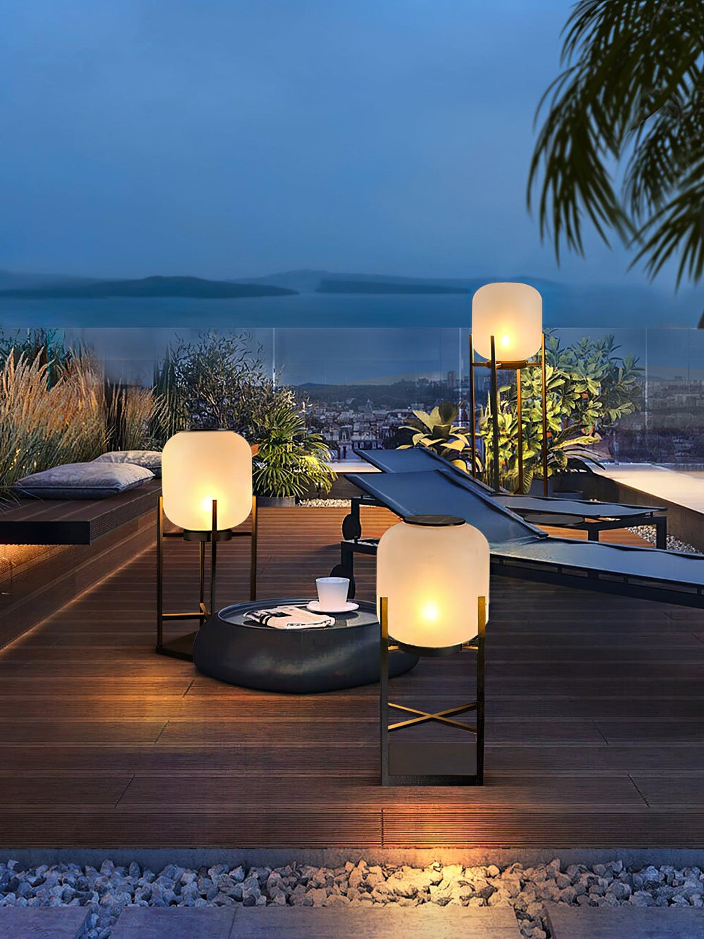 Wide view of three Clérachic lanterns lit on a deck, with a round fire bowl, a lounger and a sea view at dusk.