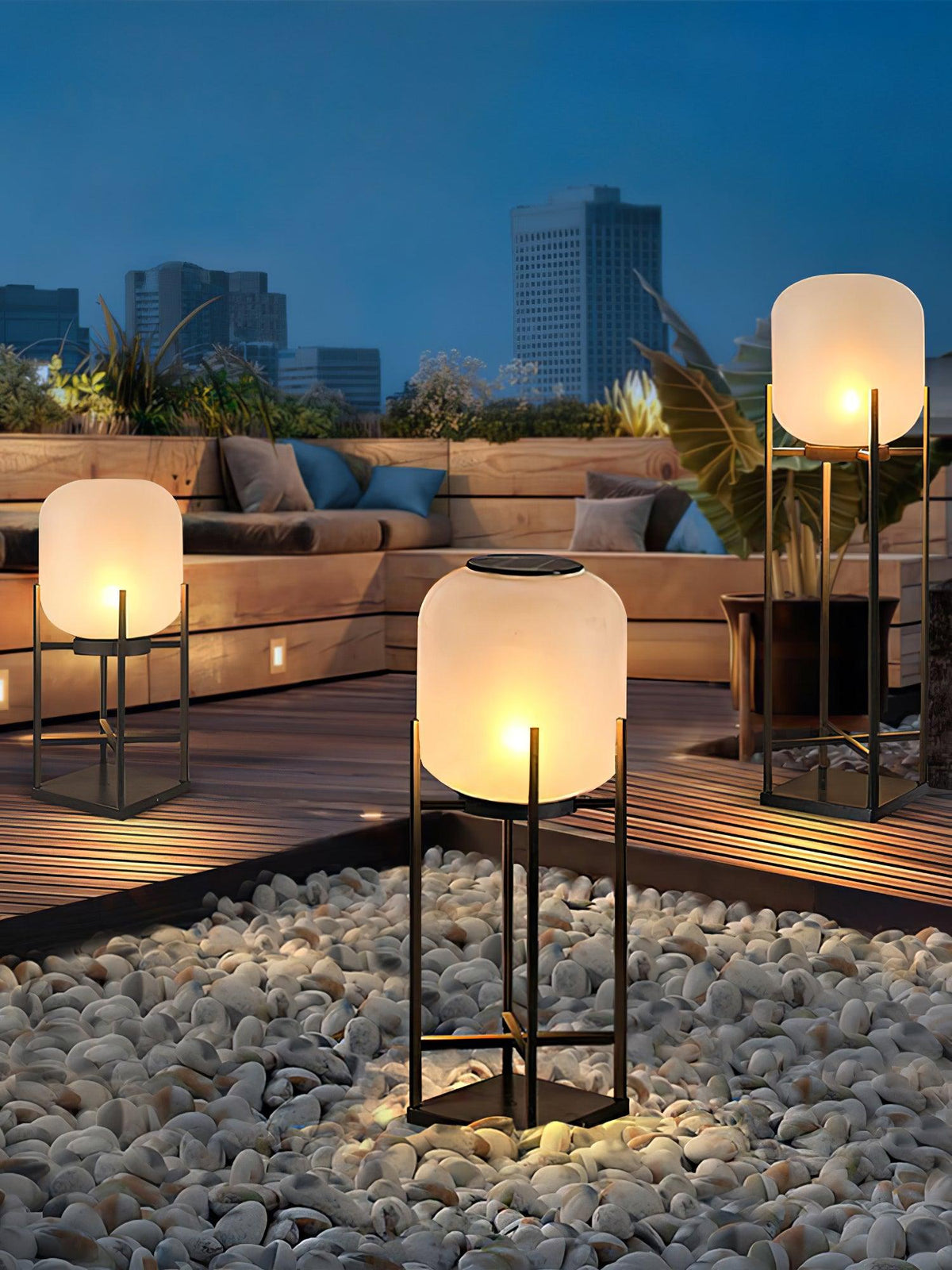 Clérachic lantern glowing in a bed of white pebbles on a roof terrace, with a sofa and city skyline in the dusk behind.