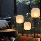 Three lit Clérachic lanterns at different heights beside a dark sofa and a round table, with woodland softly behind.