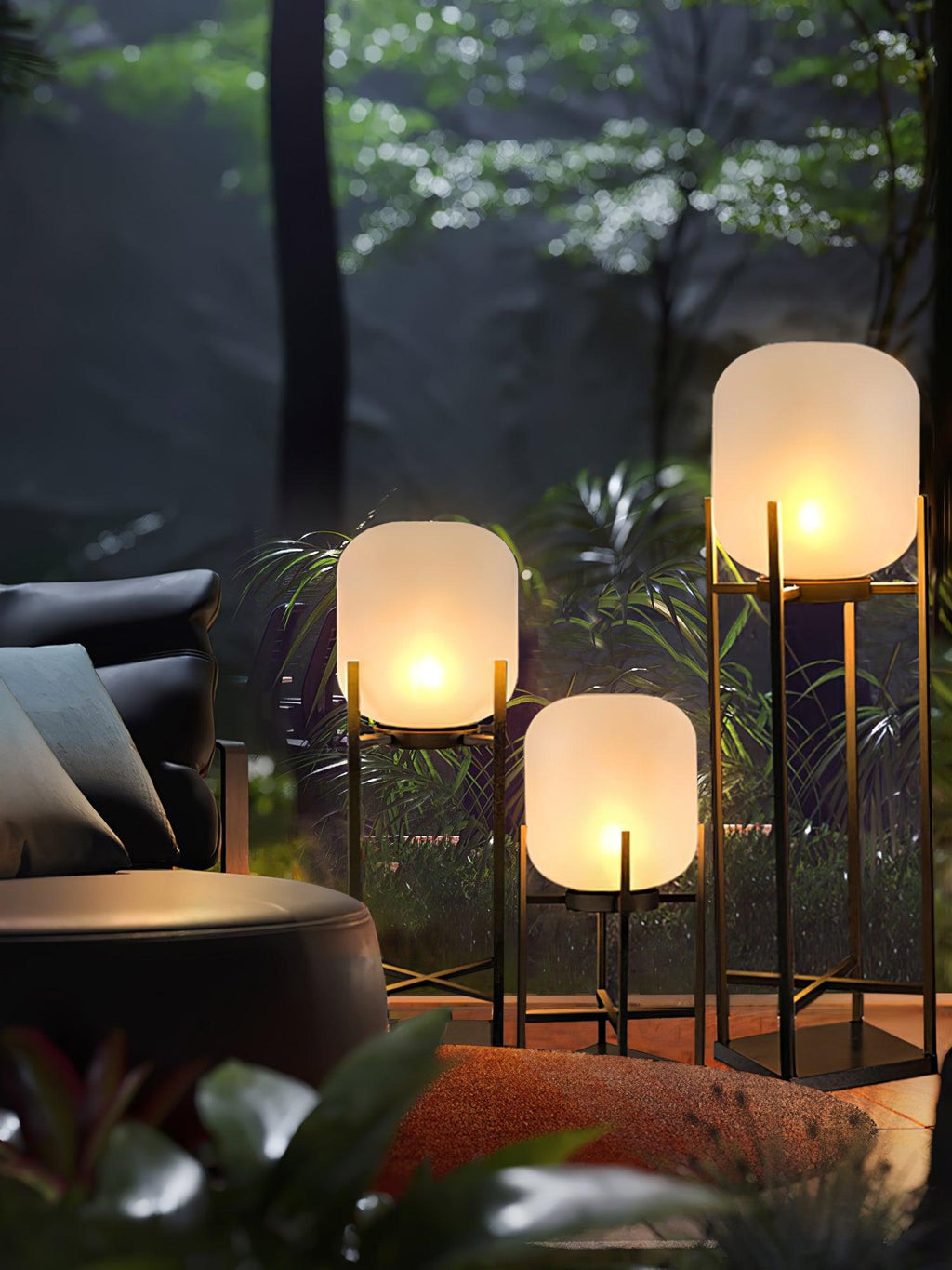 Three Clérachic lanterns glowing beside a dark outdoor sofa and a round table, with trees in the dark behind them.