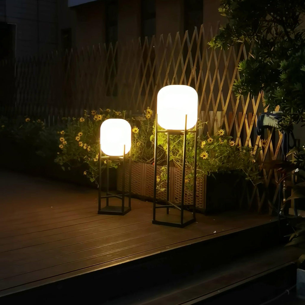 Two Clérachic lanterns of different heights lit beside a trellis fence and planters of yellow flowers at night.