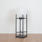 Slightly angled view of this Clérachic lantern unlit on a wooden floor, with its white shade and charcoal four-post frame.