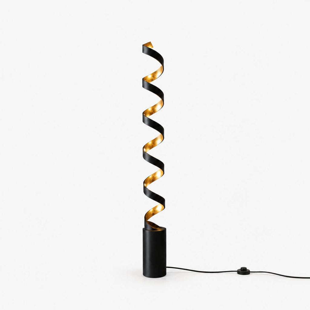 Slim black spiral floor lamp from Clérachic with a gold interior, standing on a plain white backdrop with its foot-switch cord on the floor.