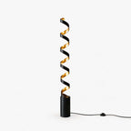 Slim black spiral floor lamp from Clérachic with a gold interior, standing on a plain white backdrop with its foot-switch cord on the floor.