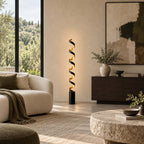 A Clérachic spiral floor lamp glowing warm beside a dark sideboard, with a cream sofa, a vase of olive sprigs and a large abstract canvas behind.