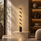 The gold inner face of this Clérachic spiral floor lamp lit up on a pale stone plinth, with ceramic vases on warm shelves behind.