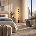 A Clérachic spiral floor lamp lit next to a bed with linen curtains, a pale armchair and a tall window looking out over a city.