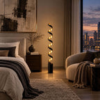 Evening view of a bedroom with this Clérachic spiral floor lamp lit beside a dark wooden nightstand and a window onto the city lights.