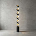 This Clérachic spiral floor lamp lit in front of a bare concrete wall, its black cylinder base standing on a smooth pale floor.