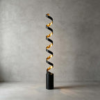 This Clérachic spiral floor lamp lit in front of a bare concrete wall, its black cylinder base standing on a smooth pale floor.