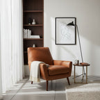 A Clérachic black floor lamp beside a tan leather armchair and a brass side table, in front of a walnut shelving unit.