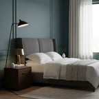 In a bedroom with blue-grey panelled walls, this Clérachic floor lamp reaches over a grey upholstered bed.