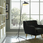 The Clérachic floor lamp's angled shade glows warm above a dark armchair, in front of a tall window.