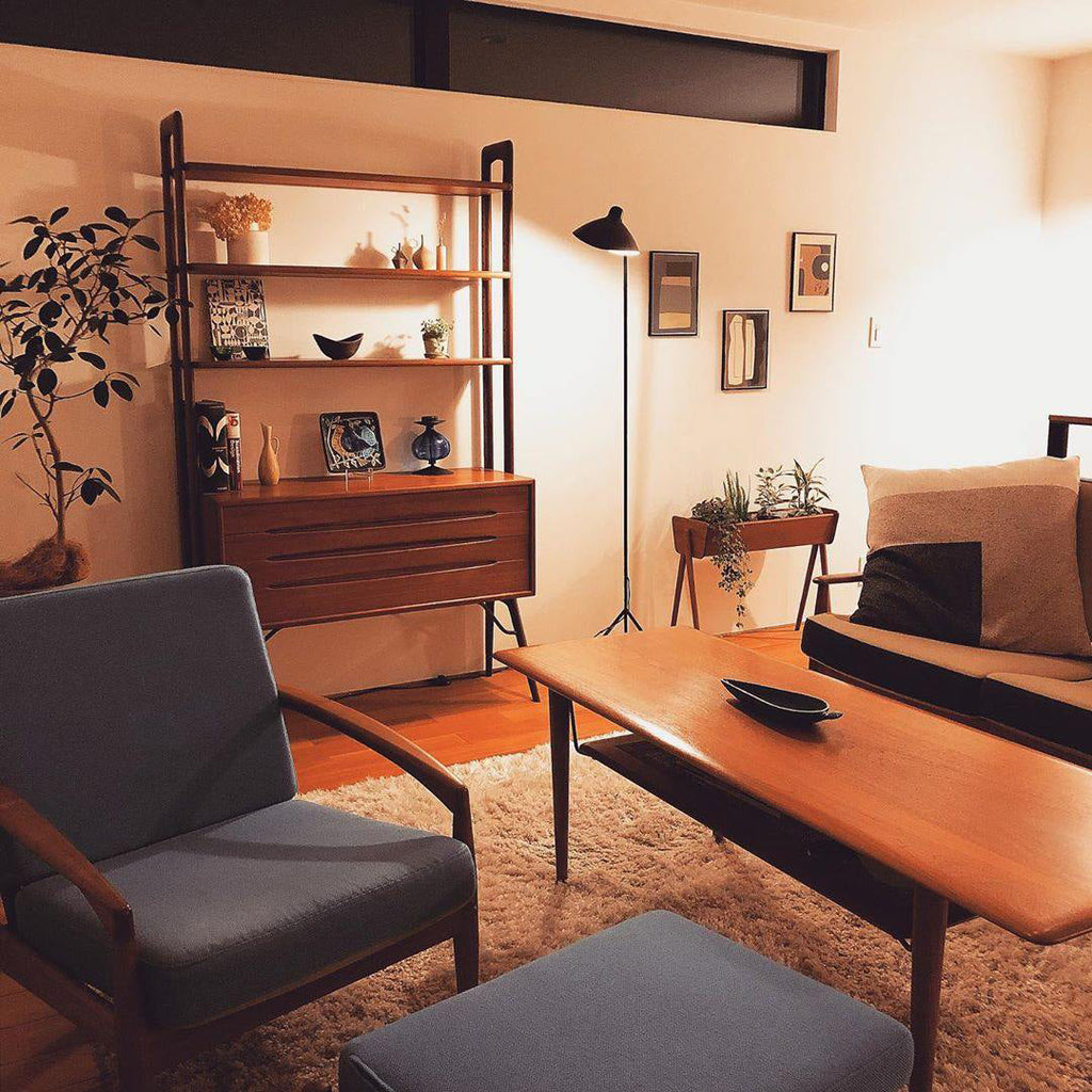 A Clérachic floor lamp stands in a retro living room with a teak shelving unit, a low timber table and a grey armchair.