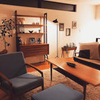 A Clérachic floor lamp stands in a retro living room with a teak shelving unit, a low timber table and a grey armchair.