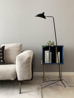 Beside a cream armchair and a small blue shelf, this Clérachic floor lamp rises from a tripod base against a grey wall.